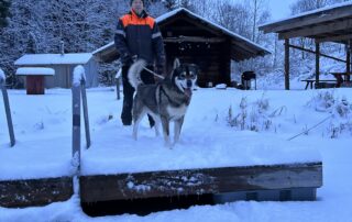 Sauna master Silver standing with his dog in front of a traditional smoke sauna, surrounded by a snowy winter landscape